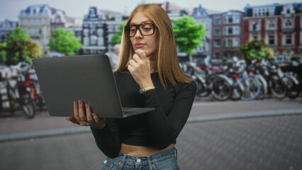 Fototapeta premium Woman holding laptop touching chin with hand on a busy street beside rows of parked bicycles and historic buildings; thoughtful focus.