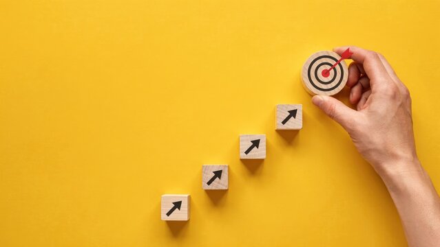A hand placing a target icon at the top of wooden blocks with upward arrows on a yellow background. Business strategy and career growth concept for corporate presentations.