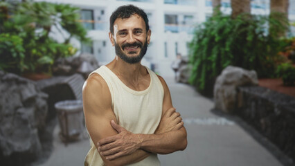 Fototapeta premium Man standing with arms crossed showing muscular forearms and smile, wearing sleeveless top in a hotel building courtyard; confidence.
