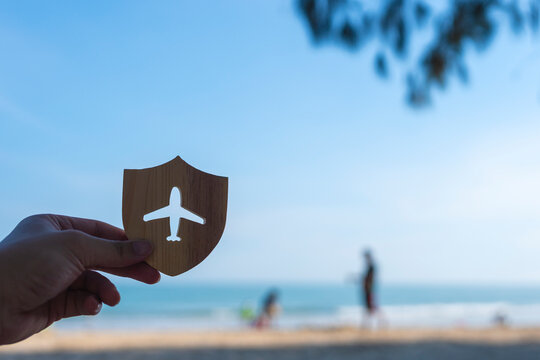Travel insurance and flight protection concept with hand holding wooden shield with airplane cutout against tropical sea beach and blue sky for vacation safety and trip cancellation coverage.
