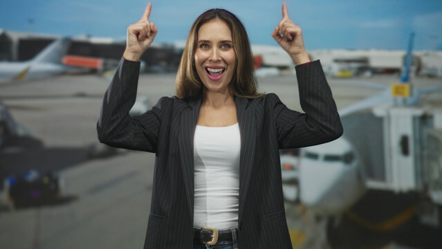 Woman pointing index fingers upward at airport jetway, smiling with raised arms and blazer, white top visible; joy optimism.