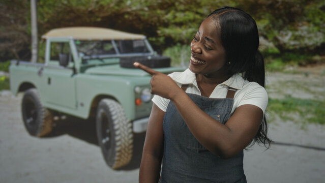 Woman wearing denim apron points finger with right hand and smiles beside mint green vintage truck in studio; warm joy.