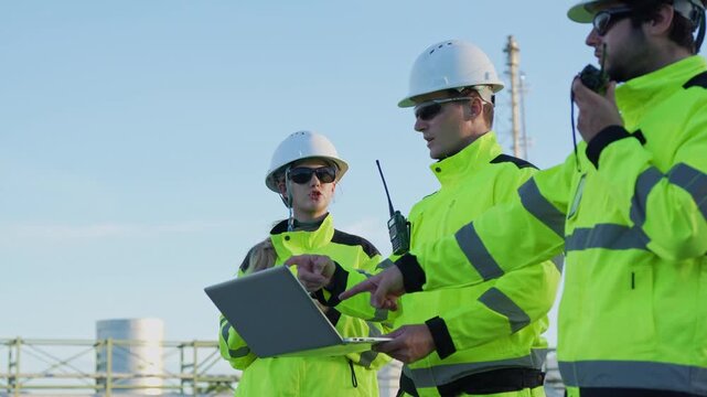 oil refinery engineers using laptop and walkie talkie outdoors, petroleum plant team working with laptop at energy facility, oil and gas engineers checking data on laptop at refinery plant