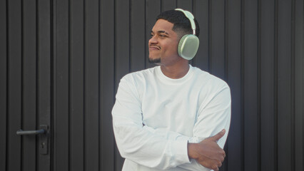 Naklejka premium Man wearing green headphones smiling with arms crossed while looking aside, standing by a black metal building door; urban calm contentment.