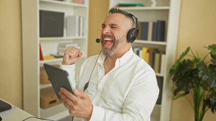 Fototapeta premium Middle age hispanic man call center agent wearing headset holds tablet, raises fist and smiles while seated in an office building with bookshelf visible; joy celebration.