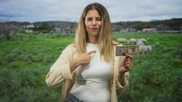 Woman holds a us fifty dollar banknote between fingers with hand on hip, posing in a forest meadow with grazing sheep and distant hills; confidence.