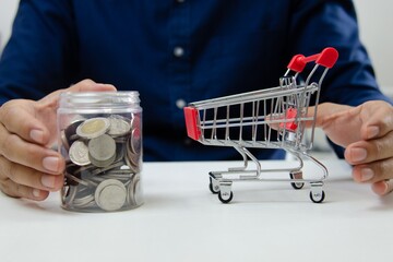 Person holding a glass jar filled with coins next to a miniature shopping cart on a table shopping currency © mrzn