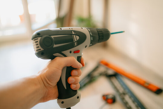 Hand holding an electric drill with bit poised to drill into a wall, tv bracket and tools blurred in background diy home renovation and mounting in progress