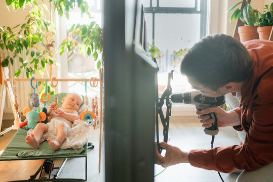 Young father carefully installing a television onto a wall bracket using a drill, focusing on the task while his newborn baby observes from a nearby bouncer in a brightly lit living room