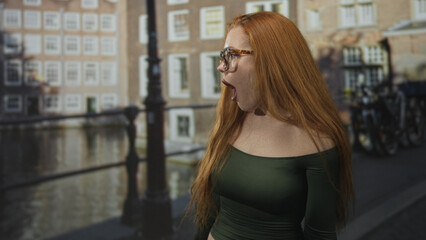 Fototapeta premium Woman with open mouth and palms up shrug on canal street by bridge railing and parked bicycles, wearing glasses and green off shoulder top; surprise wonder.
