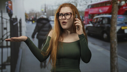 Fototapeta premium Redheaded woman on a street holding a smartphone to her ear with palm raised in an exasperated shrug, wearing glasses and green off shoulder top; frustration confrontation.