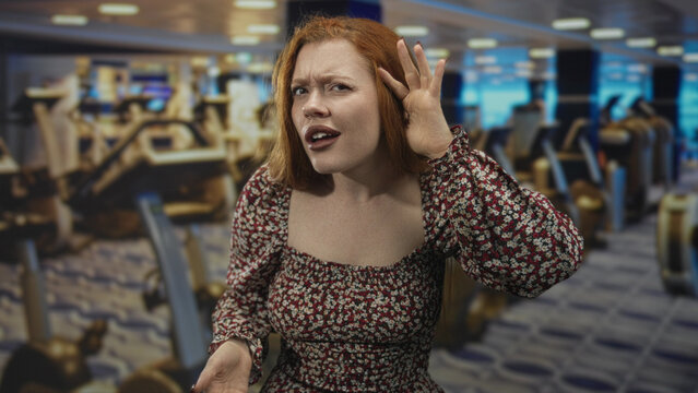 Redhead woman cups ear and points finger toward camera amid gym cardio machines in building, leaning forward with puzzled frown and raised hand; frustration confrontation.
