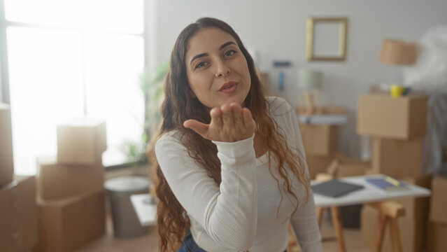 Woman surrounded by moving boxes blows a kiss with her hand reaching forward in a building; happiness new beginning.