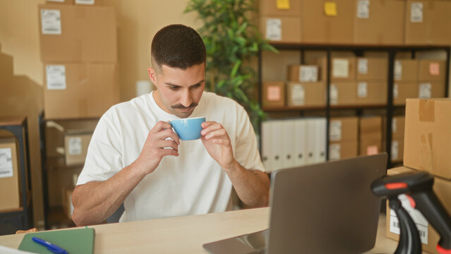 Man holding a mug while working on a laptop and checking packages in a small business building; quiet concentration.