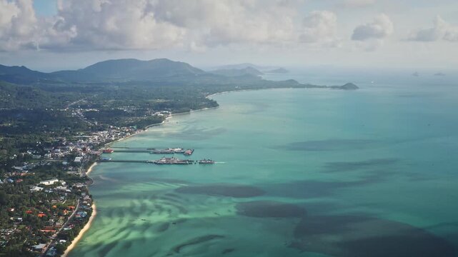 Panoramic Samui tropical island landscape. Nathon town coastline and ferry piers connecting to the mainland. Clear turquoise ocean water and coastal village houses. Aerial view drone flight zoom out