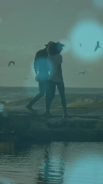 Blue spots of light floating against african american couple walking on the rocks near the sea