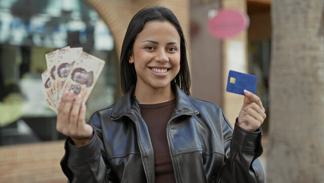 Young hispanic woman smiling while holding mexican pesos fan and a blue creditcard in front of shops on street; financial confidence.