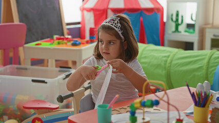 Girl cuts paper with scissors at a play table inside a building wearing a tiara and using hands to snip shapes  concentration creative play. © Krakenimages.com