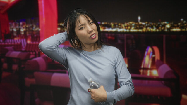 Woman young beautiful holding a water bottle, hand to mouth gesture with elbow tucked across torso in rooftop building lounge overlooking city lights at night; pensive solitude.