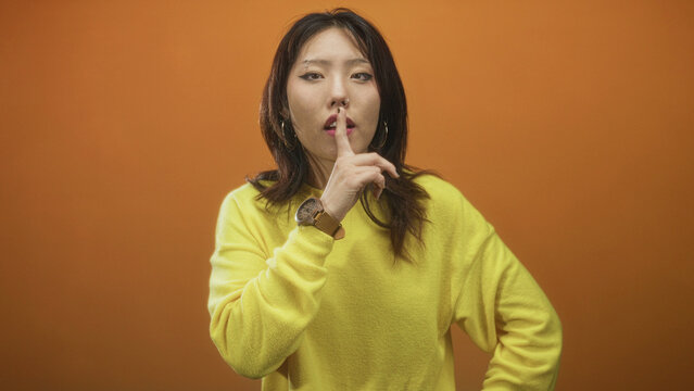 Woman in yellow sweater with wristwatch and nose piercing presses finger to lips as hush gesture in studio; silence secrecy.