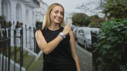 Fototapeta premium Woman smiling and pointing finger at a parked car on a residential street, wearing sleeveless black top, watch and bracelets by iron railing and townhouse steps; cheerful greeting.