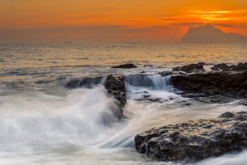 Fototapeta premium Sunbeams breaking through dark storm clouds over the sea with distant ships and rocky shore at sunset in Miono Cantabria