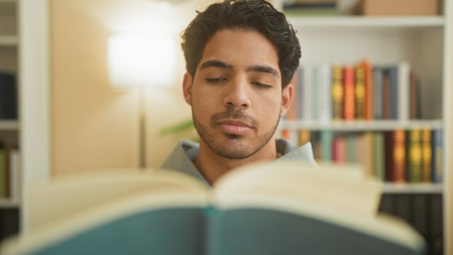 Young man reading a large open book, eyes lowered and holding pages, seated in a study building with bookshelves and a lamp visible; quiet concentration.