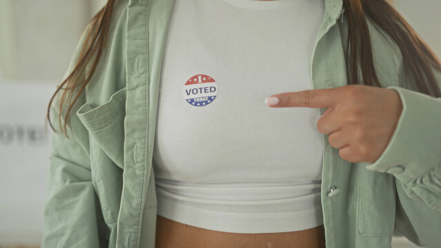 Woman pointing finger to voted sticker on white shirt inside polling building, cropped torso shows hand gesture and casual green jacket layers; civic pride.
