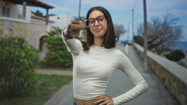 Young woman in white shirt flips middle finger to camera on a seaside street promenade, hand extended and smirking; rebellion defiance.