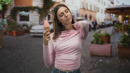 Fototapeta premium Woman holding an ice cream cone, hand on neck and slight grimace, wearing pink top and jeans while standing on a cobbled street; discomfort.