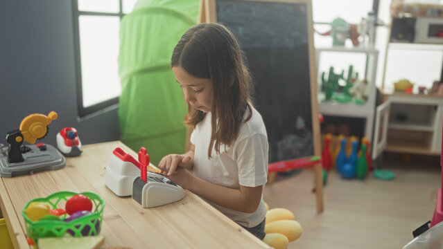 Little girl pressing buttons on a toy cash register with fingers and focused gaze in a building playroom; concentration imagination play.