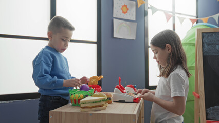 Naklejka premium Girl and boy play with toy food and a cash register on a wooden table inside a building; playful cooperation.
