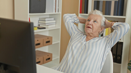 Fototapeta premium Woman senior removing glasses and holding forehead at computer beside bookshelves in an office building, stretching between tasks; fatigue.