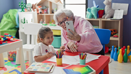 Fototapeta premium Woman holding smartphone taking selfie with little girl at colorful preschool classroom table showing drawings and colored pencils and toys on shelves; generational bonding joy.