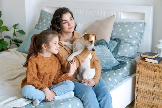 Young caucasian mom and her daughter relaxing with a jack russell terrier on a bed in a bright bedroom, ideal for home lifestyle, slow morning and pet content
