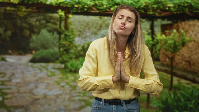 Woman with hands clasped in prayer and puckered lips pleading in a sunlit stone courtyard, wearing yellow shirt and jeans; pleading apology.