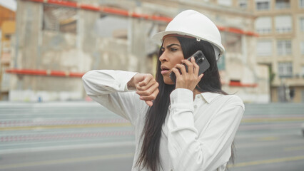 Fototapeta premium Woman architect in hardhat on building site talks on smartphone to ear while checking wristwatch and studying plans in hand; deadline urgency.
