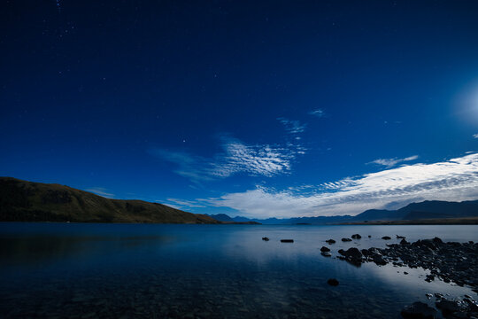 Starry night sky and moonlight over the calm water of Lake Tekapo with mountain silhouettes, Mackenzie District, South Island, New Zealand