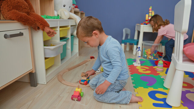 Boy kneeling on floor builds wooden train track and pushes a toy car beside foam puzzle mat and storage shelves with stuffed animals in building; playful childhood joy.