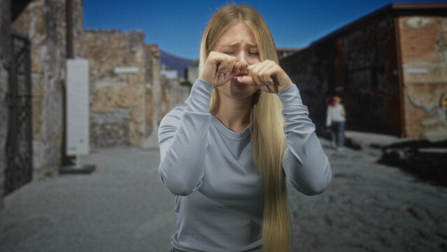 Young blonde woman with long hair rubbing hands at eyes on street near ruins and stone building wearing long sleeve sweater; sadness.
