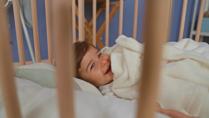 Boy lying in pale wooden crib clutching soft fleece blanket, hand near mouth, gaze sideways toward room window in building  cozy calm. © Krakenimages.com
