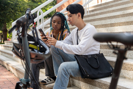 Urban Commute: Two young adults, a man and a black woman, take a break on concrete stairs beside a foldable bicycle, engrossed in their smartphones. Bike to work.