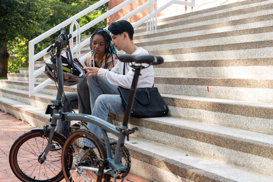 Urban Commute: Two young adults, a man and a black woman, take a break on concrete stairs beside a foldable bicycle, engrossed in their smartphones. Bike to work.