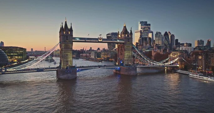 London iconic Tower Bridge spanning the River Thames, illuminated against a colorful sunset sky, with the modern City of London skyline featuring Gherkin and Shard in the background. Aerial panorama