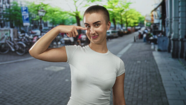 Woman with short hair points finger to cheek and smiles, wearing a white t-shirt on a city street with parked bicycles and cars; playful.