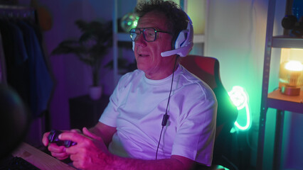 Man in headset holds game controller, hands gripping joystick while seated in studio with neon controller sign and shelving  senior gaming concentration. © Krakenimages.com