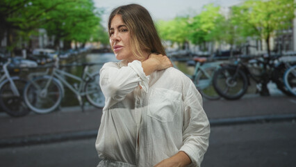 Fototapeta premium Woman with hand on neck rubbing neck in a street by a canal lined with parked bicycles, eyes closed and wincing; pain tension vulnerability.