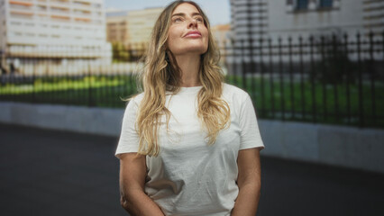 Fototapeta premium Woman in white t shirt gazes upward with closed eyes and slight smile, forearms exposed, standing on street by iron fence and city buildings; serene reflection.