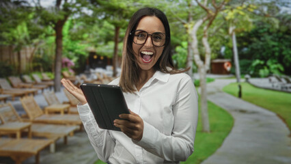 Woman holding tablet with both hands, smiling and tapping screen while standing beside wooden lounge chairs on a paved path near a building  joyful connection. © Krakenimages.com
