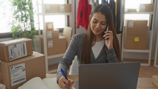 Woman holds smartphone to ear and writes with pen beside stacked shipping boxes and laptop in building; focused entrepreneurship.